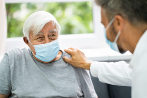 A male doctor places a comforting hand the shoulder of an older male patient during a medical visit. Both men are wearing surgical masks.