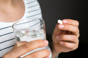 A close-up image of a woman in a striped shirt holding a pill in one hand and a glass of water in the other.