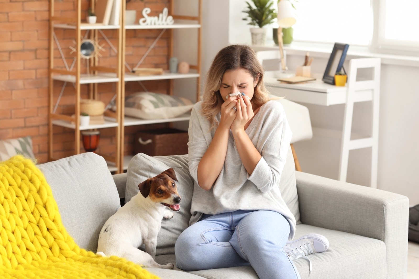 A woman sitting on the couch next to her dog, blowing her nose.