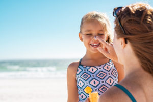 A woman applies sunscreen to the nose of a female child who is smiling at the beach.