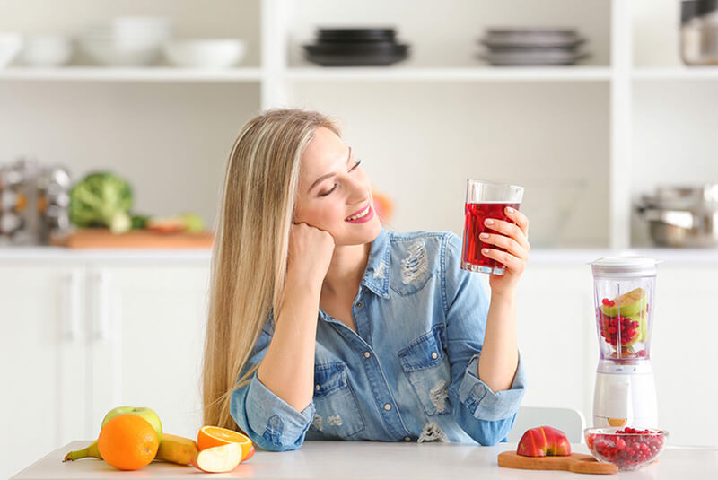 A blonde woman sitting at a countertop in her kitchen next to a blender and fruit, smiling at a glass of fresh cranberry juice in her hand.