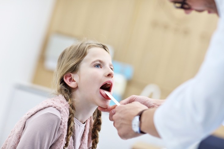 A doctor in a white lab coat is examining a young girl’s throat in an exam room using a tongue depressor.