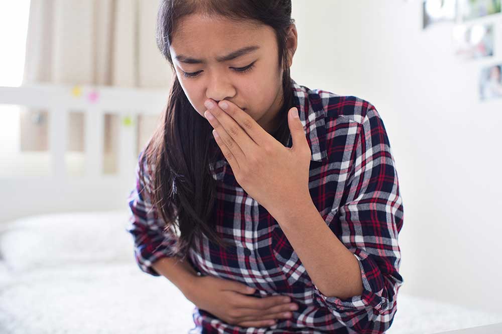 A young girl in a plaid shirt sitting on her bed holding her stomach with her hand to her mouth in pain.
